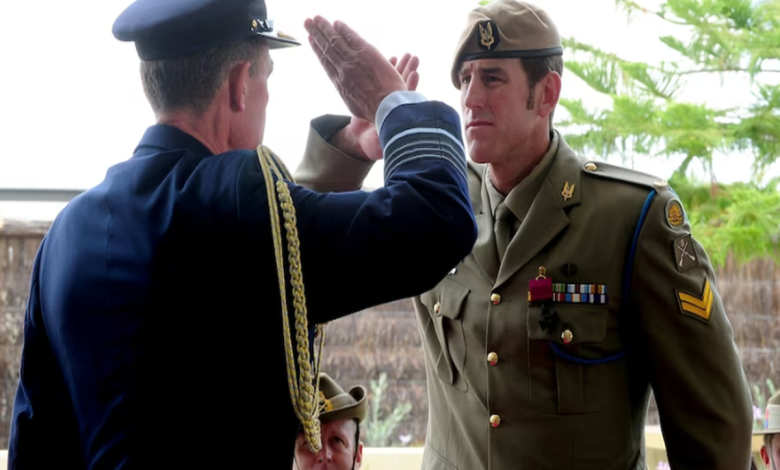 군복 차림의 벤 로버츠-스미스와 법원 또는 경찰 호송 장면을 연상시키는 이미지, 전쟁 영웅의 추락을 상징